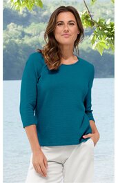 Türkisfarbenes Langarmshirt ohne Muster, Rundhalsausschnitt, gerade geschnitten, getragen von stehender Frau mit linker Hand in Hosentasche