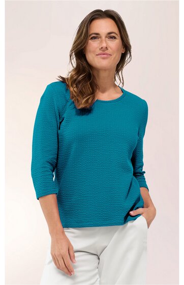 Damenshirt in petrolblau, feines Rippmuster, Rundhalsausschnitt, 3/4-lange Ärmel, gerade geschnitten, getragen von stehender Frau mit rechter Hand in Hosentasche