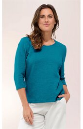 Damenshirt in petrolblau, feines Rippmuster, Rundhalsausschnitt, 3/4-lange Ärmel, gerade geschnitten, getragen von stehender Frau mit rechter Hand in Hosentasche