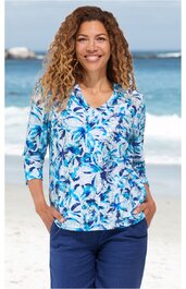Langarmshirt mit V-Ausschnitt, weiß mit blauem Blumenmuster, locker sitzend, getragen von lächelnder Frau mit lockigen Haaren, Hände in Hosentaschen stehend vor Strandkulisse