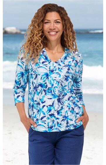 Langarmshirt mit V-Ausschnitt, weiß mit blauem Blumenmuster, locker sitzend, getragen von lächelnder Frau mit lockigen Haaren, Hände in Hosentaschen stehend vor Strandkulisse