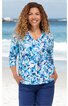 Langarmshirt mit V-Ausschnitt, weiß mit blauem Blumenmuster, locker sitzend, getragen von lächelnder Frau mit lockigen Haaren, Hände in Hosentaschen stehend vor Strandkulisse
