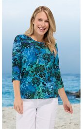 Damenoberteil, blau mit grünem Blumenmuster, rundhalsausschnitt, 3/4-lange Ärmel, locker sitzend, getragen von stehender Frau vor Strandhintergrund