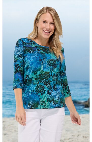 Damenoberteil, blau mit grünem Blumenmuster, rundhalsausschnitt, 3/4-lange Ärmel, locker sitzend, getragen von stehender Frau vor Strandhintergrund