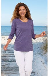 Lila Pullover mit Rundhals, drei Viertel Ärmel, schlichte Form, getragen von stehender Frau am Strand, weiße Hose, entspannte Pose