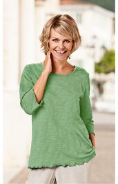 Hellgrünes Langarmshirt ohne Muster, Rundhalsausschnitt, gerader Schnitt, leicht strukturierter Stoff, gewellte Saumkanten, getragen von lächelnder stehender Frau mit kurzer blonder Frisur