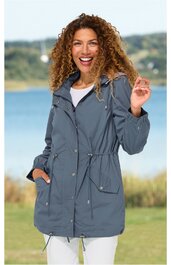 Graue lange Jacke mit Tunnelzug an Taille und Saum, Stehkragen mit Kapuze, Reißverschluss und Druckknöpfen, aufgesetzte Taschen mit Klappen, getragen von lächelnder Frau, stehende Pose, lockere Passform