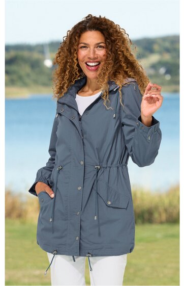 Graue lange Jacke mit Tunnelzug an Taille und Saum, Stehkragen mit Kapuze, Reißverschluss und Druckknöpfen, aufgesetzte Taschen mit Klappen, getragen von lächelnder Frau, stehende Pose, lockere Passform