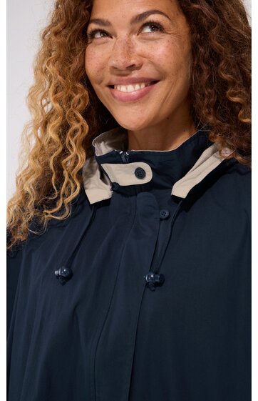Dunkelblauer Regenponcho mit beigem Kragenbesatz, Knöpfen am Kragen, Kordelzug mit Stoppern an der Brust, getragen von lächelnder Frau mit lockigen Haaren