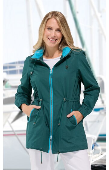 Dunkelgrüne Damenjacke mit türkisfarbenem Stehkragen, durchgehendem Reißverschluss, Tunnelzug in der Taille, langen Ärmeln, zwei seitlichen Taschen, getragen von lächelnder stehender Frau vor Wassersportkulisse
