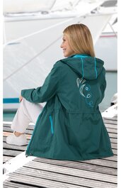 Dunkelgrüne Jacke mit türkisfarbenen Kordeln, Kapuze, großer Rückenapplikation mit geschwungenen Linien und Punkten, taillierter Schnitt, getragen von sitzender Person, weiße Hose