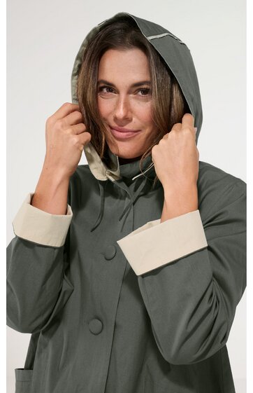 Olivgrüner Regenmantel mit beigen Ärmelumschlägen, Kapuze mit Kordelzug, Druckknöpfe vorne, getragen von frontal sichtbarer Frau, lange Ärmel