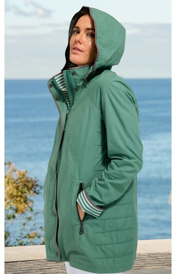 Grüne Kapuzenjacke mit Reißverschluss, seitlichen Taschen, gesteppten Details am unteren Teil, gestreiften Bündchen, getragen von stehender Frau vor Meereskulisse