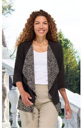 Schwarze offene Strickjacke mit wehendem, floral gemustertem Einsatz in Weiß-Schwarz, dreiviertellange Ärmel, getragen von lächelnder Frau mit lockigem Haar, weiße Bluse, beige Hose