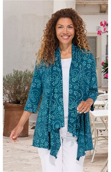 Türkisfarbene Strickjacke mit weißem, kreisförmigem Muster, offene Vorderseite, dreiviertellange Ärmel, asymmetrischer Saum, getragen von stehender Frau über weißem Top und weißer Hose