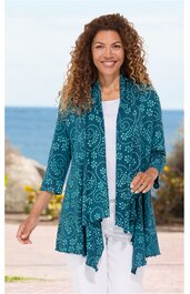 Türkise Strickjacke mit weißen kreisförmigen Mustern, offene Vorderseite mit asymmetrischem Saum, 3/4-Ärmel, getragen von lächelnder Frau in lässiger Pose vor Meereskulisse