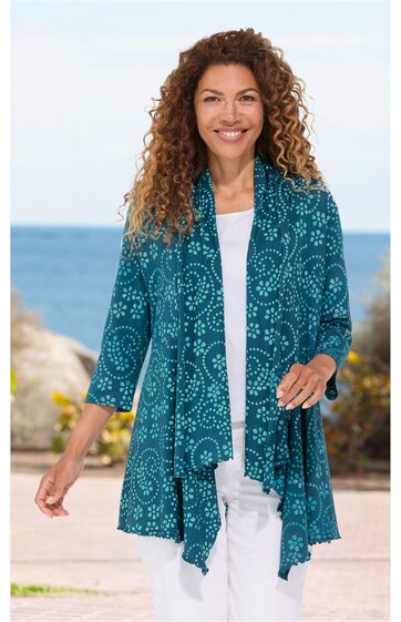 Türkise Strickjacke mit weißen kreisförmigen Mustern, offene Vorderseite mit asymmetrischem Saum, 3/4-Ärmel, getragen von lächelnder Frau in lässiger Pose vor Meereskulisse