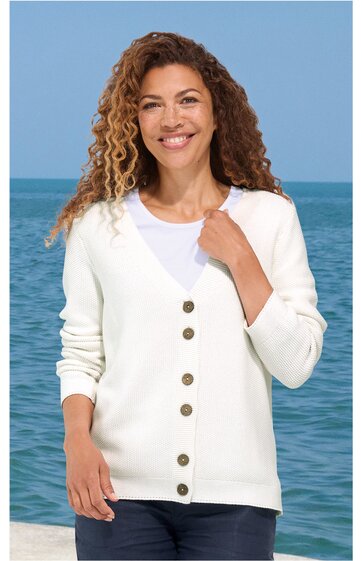 Weißer Damenstrickcardigan mit V-Ausschnitt und sechs braunen Knöpfen, langärmlig, gerade geschnitten, getragen von lächelnder Frau vor Meereskulisse