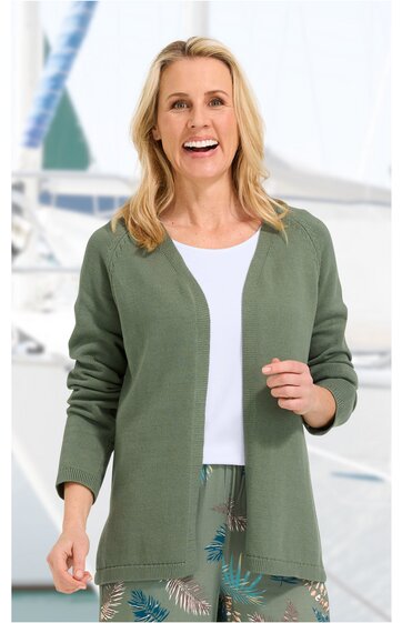 Olivgrüne offene Strickjacke ohne Verschluss, lange Ärmel, gerade geschnitten, getragen von lächelnder stehender Frau mit weißen T-Shirt und gemusterten grünen Hosen, Hintergrund mit Segelboot
