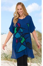 Blaues Tunika-Kleid mit abstraktem, mehrfarbigem Muster in Grün, Lila und Schwarz, V-Ausschnitt, kurze Ärmel, asymmetrischer Saum, getragen von stehender Frau am Strand