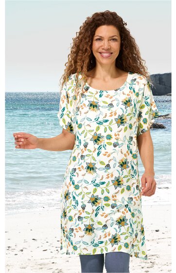 Weißes Kleid mit floralem Muster in Grün und Gelb, rundem Ausschnitt, kurze Ärmel, knielang, gerade geschnitten, getragen von lächelnder Frau, stehend am Strand