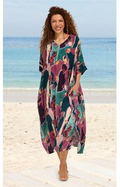 Maxikleid in dunklem Türkisgrün mit buntem abstraktem Muster in Rosa, Lila und Orange, V-Ausschnitt mit Knopfleiste, kurze weite Ärmel, Seitentaschen, lockere weite Passform, getragen von stehender Frau am Strand
