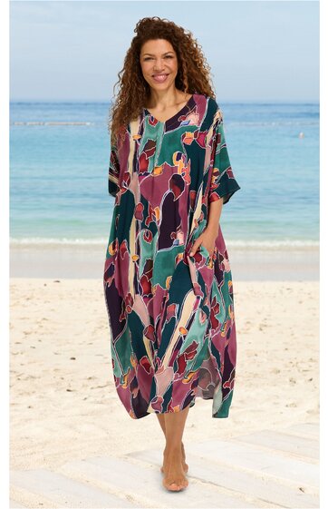 Maxikleid in dunklem Türkisgrün mit buntem abstraktem Muster in Rosa, Lila und Orange, V-Ausschnitt mit Knopfleiste, kurze weite Ärmel, Seitentaschen, lockere weite Passform, getragen von stehender Frau am Strand
