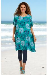 Knielanger, locker geschnittener Tunika-Kleid in türkis mit abstraktem Kreismuster in verschiedenen Blautönen, rundem Ausschnitt, kurze Raglanärmel, getragen von stehender Frau am Strand, kombiniert mit dunkelblauen Leggings und türkisfarbenen Sandal