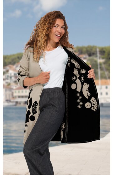Beige Cardigan mit schwarzen grafischen Mustern, offen getragen von lächelnder stehender Frau mit lockigen Haaren, weiße T-Shirt, graue Hose, Innenfutter schwarz mit beige gemustert