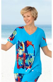 Blaues T-Shirt mit buntem abstraktem Muster in Rot, Gelb, Weiß und Schwarz, V-Ausschnitt, kurze Ärmel, getragen von lächelnder stehender Frau vor Meereskulisse
