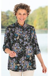 Schwarze Bluse mit Blumenmuster in Blau, Lila und Beige, Stehkragen, verdeckte Knopfleiste, 3/4-Ärmel, locker geschnitten, getragen von stehender Frau vor unscharfem Naturhintergrund