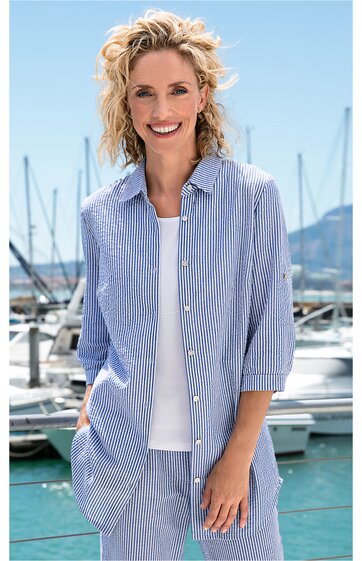 Blau-weiß gestreifte Hemdbluse mit langem Schnitt, Kragen, Knopfleiste, weite Passform, 3/4-Ärmel, getragen von stehender Frau vor Hafen mit Booten