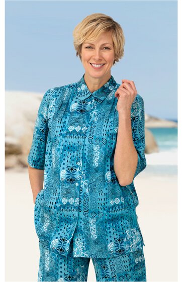 Blaues Hemd mit abstraktem Muster in Dunkel- und Hellblau, kurzärmlig, mit klassischem Kragen, durchgehender Knopfleiste, locker sitzend, getragen von stehender Frau am Meer
