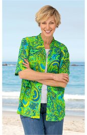 Grüne Bluse mit Paisley-Muster in Gelb und Blau, halblange Ärmel, V-Ausschnitt mit Umlegekragen, offen getragen über weißem T-Shirt, getragen von lächelnder Frau mit verschränkten Armen vor Meereskulisse