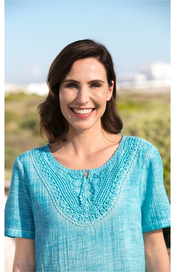 Türkisfarbenes Oberteil mit kurzem Ärmel, strukturierte Häkelspitze am rundem Ausschnitt, lockere Passform, getragen von lächelnder Frau mit schulterlangen braunen Haaren, stehende Haltung