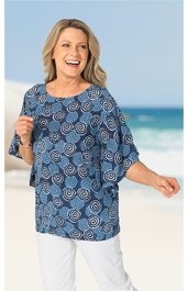 Damenbluse in Blau mit weißem Spiralmuster, rundem Ausschnitt, kurze weite Ärmel, lockere Passform, getragen von lächelnder stehender Frau vor Strandhintergrund