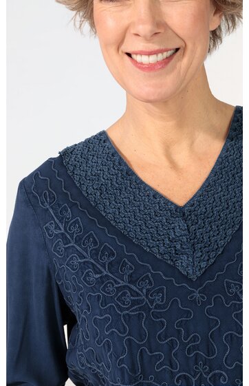 Dunkelblaues Oberteil mit V-Ausschnitt, umlaufendem Muster aus gehäkelter Borte am Kragen, dekorative Stickerei auf Vorderseite, langärmelig, getragen von stehender Person