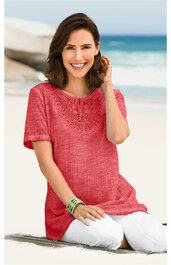 Rotes Kurzarmshirt mit gestricktem Muster am Ausschnitt, Rundhalsausschnitt, lockere Passform, getragen von kniender Frau mit weißen Hosen und Armbändern vor Meereshintergrund