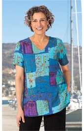 Kurzärmeliges Freizeitshirt in Blau- und Türkistönen mit abstraktem Muster aus Rechtecken und Punkten, V-Ausschnitt, gerade geschnitten, getragen von lächelnder Frau im Stehen mit angelegten Armen