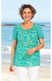 Türkisfarbenes Kurzarmshirt mit gelbem Blumenmuster, runder Ausschnitt, gerade geschnitten, getragen von stehender Frau am Strand, lockeres Oberteil