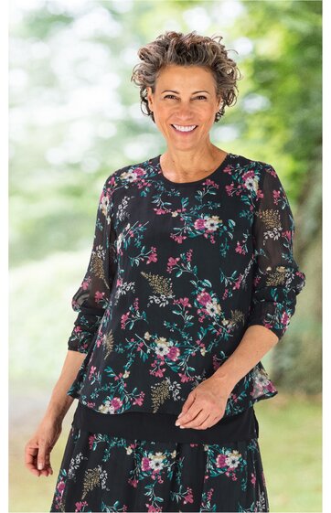 Schwarze Bluse mit buntem Blumenmuster, rundem Ausschnitt, lange transparente Ärmel mit Gummizug, locker sitzend, getragen von lächelnder Frau in aufrechter Pose
