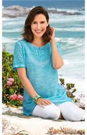 Hellblaues Damen-T-Shirt mit kurzem Arm, Rundhalsausschnitt, gehäkeltem Einsatz am oberen Vorderteil, locker sitzend, getragen von sitzender Frau am Strand, weiß, bequeme Passform