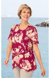Shirt in dunkelrosa mit beigefarbenem großflächigem Blumenmuster, Rundhalsausschnitt, kurze Ärmel mit Schlitzöffnung, oben überlappende Front, getragen von lächelnder Frau im Stehen vor Wasser und Felsen