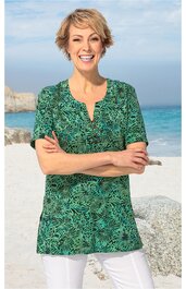 Grünes T-Shirt mit schwarzem Blattmuster, kurzer Knopfleiste am Rundhalsausschnitt, halblange Ärmel, gerade geschnitten, getragen von lächelnder stehender Frau am Strand