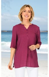 Dunkelrosafarbene Bluse mit 3/4-Ärmeln, Rundhals mit kleinem V-Ausschnitt, durchgehende Knopfleiste, locker geschnitten, getragen von stehender Frau vor Strandkulisse