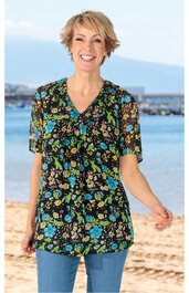 Schwarze Tunika mit buntem Blumenmuster in Blau, Grün und Gelb, V-Ausschnitt, kurze transparente Puffärmel, gerade geschnitten, getragen von lächelnder stehender Frau am Strand