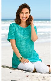 Türkisfarbenes Kurzarmshirt mit rundem Halsausschnitt, floraler Strickstruktur vorne, locker sitzend, getragen von sitzender Frau am Strand, mit weißen Hosen und mehreren Armbändern