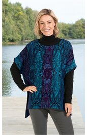 Kurzärmliges Damenoberteil, dunkelblau mit türkis und violett, abstraktes Muster, weiter Rundhalsausschnitt, kastige Form, getragen von lächelnder stehender Frau mit schwarzem Rollkragenpullover und grauer Hose