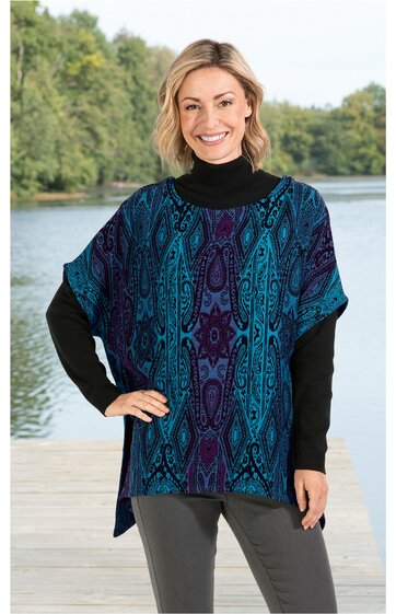 Kurzärmliges Damenoberteil, dunkelblau mit türkis und violett, abstraktes Muster, weiter Rundhalsausschnitt, kastige Form, getragen von lächelnder stehender Frau mit schwarzem Rollkragenpullover und grauer Hose