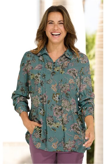Langärmlige Bluse in grünblau mit Blumenmuster, Hemdkragen, Knopfleiste, locker geschnitten, getragen von lächelnder Frau, Hände in Taschen, kombiniert mit violetten Hosen